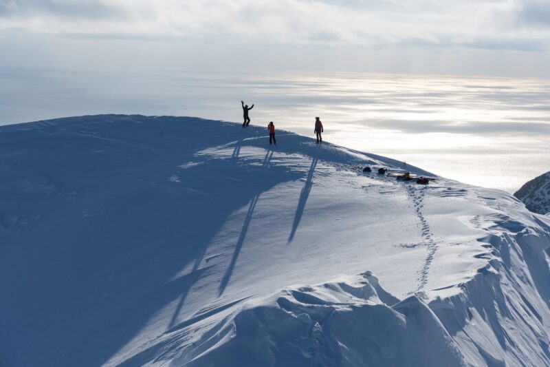 Sailboat anchored in an Arctic fjord and backcountry skiiers on top of the mountain for our Ski and sail expedition on Svalbard.