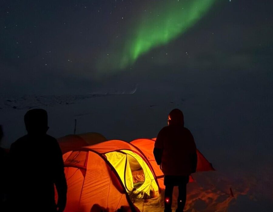 Guests in front of the orange tents in the middle of the wilderness admiring northern lights above on the Arctic Svalbard sky