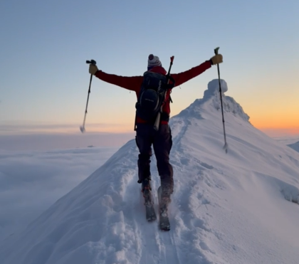 Skier standing on top of the mountain summit celebrating the vibrant orange and pink glow on the horizon during winter on Svalbard