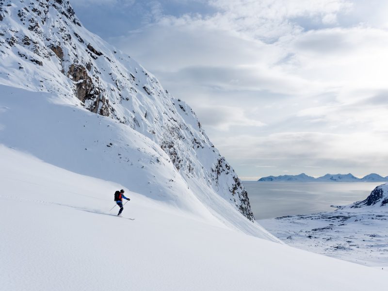 backcountry skier descending an untouched mountain slope in Svalbard with the historic expedition ship Saeftinghe waiting in the Arctic ocean below. The greatest off-pist adventure